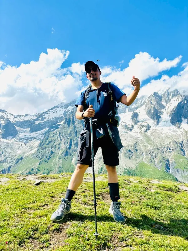 Biplab Subedi standing on a grassy ridge in the Alps with a hiking pole, snowy peaks behind him
